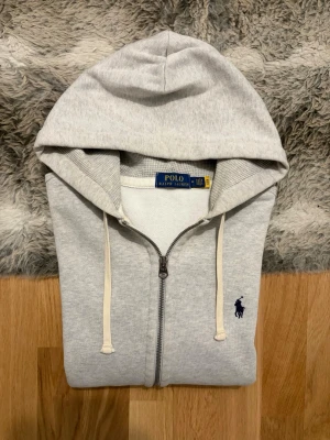 Ljusgrå Polo Ralph Lauren Zip Up Hoodie - Storlek M - Hej! 👋 Säljer en riktigt snygg ljusgrå Polo Ralph Lauren zip-up hoodie i storlek M med klassisk marinblå pony-logga. 🐎 Tröjan är i gott skick, välvårdad och känns väldigt fräsch. 100% äkta med originaltvättlappar kvar. QR-koden finns på lappen och fungerar precis som den ska för smidig verifiering ✅ Det ultimata och mest tidlösa basplagget till garderoben! Fraktar snabbt! 🚛📦💬 