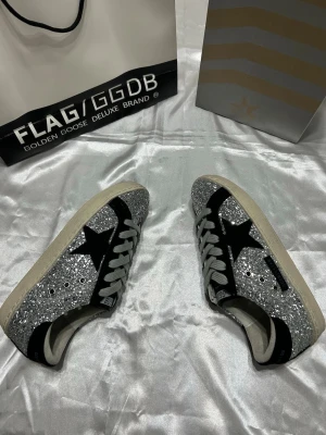 Silverglittriga Golden Goose sneakers - Snygga sneakers från Golden Goose med silverglittrig ovandel, svarta detaljer och vit sula. Skorna har den klassiska stjärnan på sidan och gråa skosnören. Perfekta för dig som vill sticka ut med en unik och trendig look.