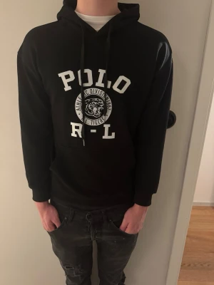 Svart hoodie från Polo Ralph Lauren - Svart hoodie från Polo Ralph Lauren med vit POLO R-L text och tigertryck på bröstet. Klassisk huva med dragsko och känguruficka framtill. Perfekt för en avslappnad och stilren look. Tillverkad i mjukt material för extra komfort. Storlek L men sitter som M