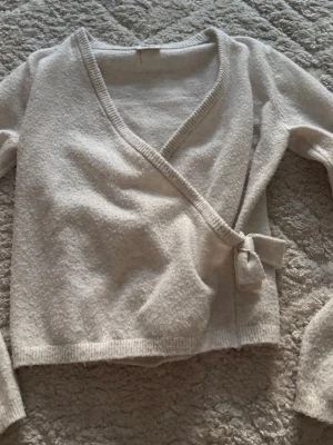 Beige omlottkofta med knytband - Mysig beige omlottkofta med knytband i sidan och ribbade kanter. Koftan har en djup V-ringning och är tillverkad i ett mjukt, stickat material. Perfekt för lager-på-lager och en chill look.