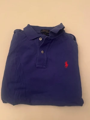 Blå pikétröja från Ralph Lauren - Klassisk blå pikétröja från Ralph Lauren med röd broderad logga på bröstet. Tröjan har krage, knappar framtill och är tillverkad i bomull. Perfekt för en stilren och avslappnad look.