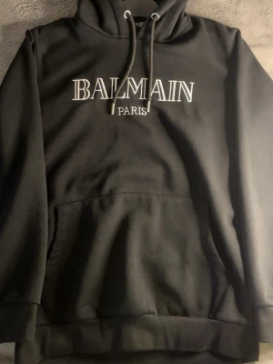 Svart hoodie från Balmain Paris - Svart hoodie från Balmain Paris med vit broderad logga på bröstet. Klassisk modell med huva och dragsnören samt stor magficka framtill. Mjuk bomullskänsla och ribbade muddar vid ärmslut och nederkant. Perfekt för en stilren och trendig look.