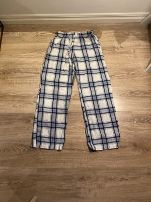 Rutiga pyjamasbyxor i blått och vitt - Snygga och bekväma pyjamasbyxor med blå och vit rutig design. Byxorna har resår i midjan och dragsko för extra komfort. Perfekta för att chilla hemma eller sova i. Tillverkade i mjukt tyg som känns skönt mot huden.