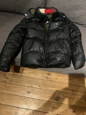 Svart pufferjacka Tommy Jeans XS  - Säljer en svart pufferjacka från Tommy Jeans i storlek XS. Jackan har hög krage, dragkedja framtill och detaljer i rött och vit på insidan. Klassisk passform och tjock vaddering som håller dig varm under kalla dagar. Perfekt för vintern! Passa på att köpa innan vintern. 