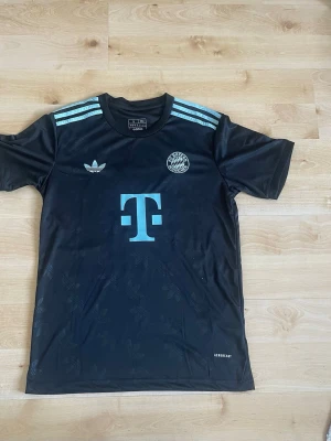 Svart Bayern München special tröja med Musiala 42 på ryggen  - Snygg svart Bayern München matchtröja från Adidas med ljusblå detaljer och tryck. Framsidan har klubbmärke, Adidas-logga och sponsortryck, medan baksidan har Musiala och nummer 42. Tillverkad i lätt och ventilerande material.