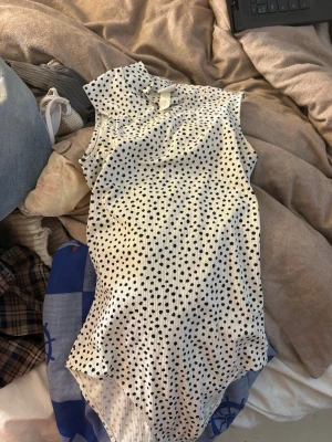 Vit prickig body från H&M XS - Man betalar frakten själv. Säljer en vit body från H&M i storlek XS med svarta prickar och hög krage. Bodyn är ärmlös och har ett lätt plisserat tyg som ger en snygg struktur. Perfekt för att skapa en trendig och stilren look.Aldrig använt packet stolek 