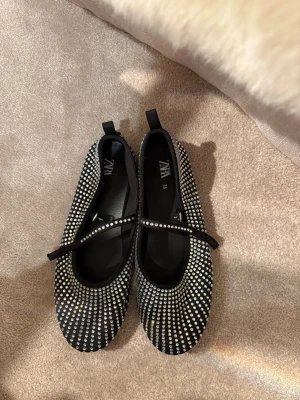 Svarta ballerinaskor med strass Zara - Snygga svarta ballerinaskor från Zara med glittrande strass över hela skon och en smal rem över foten. Perfekta för dig som vill sticka ut med lite extra bling.