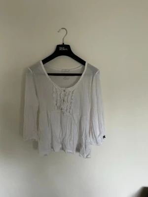 Vit blus med volang från Odd Molly - Söt vit blus från Odd Molly med volangdetaljer framtill och rund halsringning. Blusen har trekvartsärm och är tillverkad i ett lätt, tunt material som ger en avslappnad känsla. Perfekt att styla med jeans eller kjol.