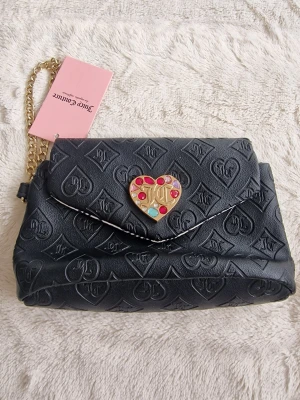 Svart Juicy Couture handväska med hjärtspänne - Cool svart handväska från Juicy Couture i präglat konstläder med JC-loggor över hela väskan. Väskan har ett guldigt hjärtformat spänne med färgglada stenar och en kedjerem i guld. Insidan är fodrad med Juicy Couture-tryck i svart på vitt tyg.