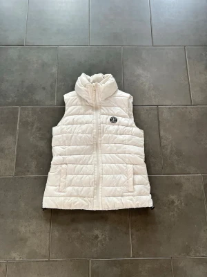 Vit dunväst  - Snygg vit dunväst med quiltat mönster och hög krage. Västen har dragkedja framtill, två fickor och en rund logotyp på bröstet. Perfekt för lager-på-lager och ger en clean look till din outfit.