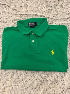 Grön pikétröja från Polo Ralph Lauren - Snygg grön pikétröja från Polo Ralph Lauren med klassisk krage och två knappar framtill. Tröjan har det ikoniska gula broderade logotypen på bröstet och är tillverkad i mjuk bomull. Perfekt för en stilren och avslappnad look.