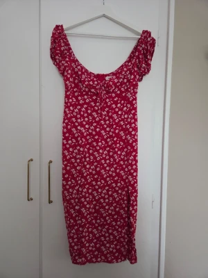 Röd blommig klänning från Nelly - Söt röd klänning från Nelly med vita blommor, korta puffärmar och knyt i ringningen. Slits vid benet.