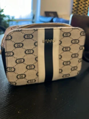 Beige axelväska från LIU JO - Snygg beige axelväska från LIU JO med svart och vit rand framtill och guldfärgad logga. Väskan har ett mönster med LIU JO-loggor över hela ytan och justerbar axelrem i svart. Tillverkad i tåligt tygmaterial med detaljer i konstläder.