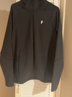 Svart hoodie från Peak Performance - Svart hoodie från Peak Performance med dragkedja framtill, två fickor och vit logga på bröstet. Huva med stor vit text på baksidan. Tillverkad i ett mjukt och stretchigt material, perfekt för en sportig och avslappnad stil.