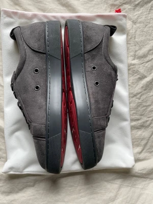 Gråa sneakers från Christian Louboutin - Snygga gråa sneakers från Christian Louboutin i mocka med ikoniska röda sulor och svarta detaljer. Skorna har en stilren design med metallfärgade öljetter och klassisk låg profil. Perfekta för dig som vill sticka ut med exklusiva detaljer.