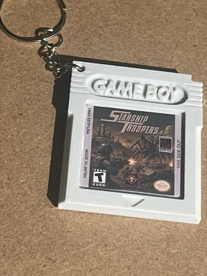 Game Boy nyckelring Starship Troopers - Säljer en unik nyckelring formad som ett Game Boy-spel med Starship Troopers-motiv. Nyckelringen är vit och har en klassisk retrostil, perfekt för dig som gillar gaming och nostalgi. Materialet är plast. Perfekt accessoar till väskan eller nyckelknippan.