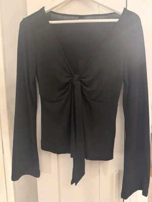 Svart långärmad topp Gina Tricot - En helt ny Snygg svart långärmad topp från Gina Tricot med djup v-ringning och knutdetalj framtill. Toppen har en figurnära passform och är gjord i ett stretchigt, mjukt material som känns skönt mot huden. Perfekt att styla med jeans eller kjol. Kan även kallas som blus då den går att använda till finare tillfällen som t.ex avslutning!