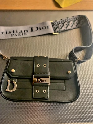 Svart handväska med bred axelrem - Snygg svart handväska med guldfärgade detaljer och ett stort D-hänge. Väskan har en bred axelrem med logotyp och ett spänne framtill med tryck. Inuti finns ett fack med dragkedja och mönstrat foder.