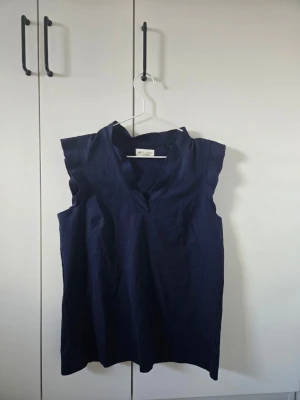 Mörkblå ärmlös blus  - Säljer en stilren mörkblå blus från Holly & Whyte med v-ringad hals och ärmlös design. Blusen har en rak passform och är gjord i ett mjukt material som känns skönt mot huden. Perfekt för dig som gillar minimalistisk och modern stil.