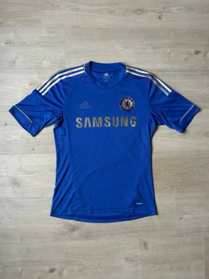 Blå Chelsea FC Hemmakit 2012/13 Fotbollströja Storlek S  - Mycket fin Chelsea fotbollströja i blå färg. Tröjan är i mycket bra skick och sparsamt använda. |  Äkta som allt annat jag säljer! ✅ |  Storlek: S | Skick: Mycket Bra Skick - Tyvärr har trycket börjat släppt litegrann på ryggen (SE SISTA BILD) | Färg: Blå | Märke: Adidas | Modell: Jeans |   Hör gärna av dig vid frågor så svarar jag så fort jag kan! 😁   (SKU - ?)