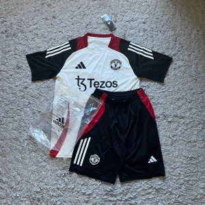 Manchester United [FAST PRIS] - Säljer ett Manchester United bortaställ från Adidas med vit tröja och svarta ärmar, röda detaljer och klubbmärke på bröstet. Matchande svarta shorts med röda inslag och klubbmärke. Materialet är lätt och andas, perfekt för fotbollsträning.