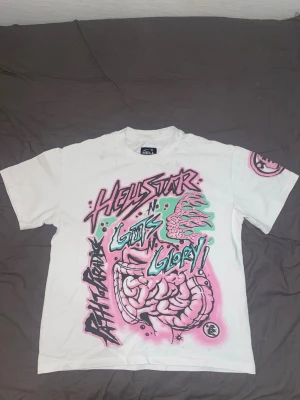 Vit Hellstar t-shirt med grafiskt tryck - Vit t-shirt från Hellstar med stort grafiskt tryck i rosa, grönt och svart både fram och bak. Trycket har text och tecknade motiv med streetwear-vibe. Rund halsringning och korta ärmar. Perfekt för dig som gillar unika statement-plagg. Obs! Defekter på bilderna