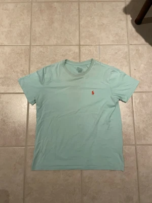Ljusblå t-shirt från Ralph Lauren - Snygg ljusblå t-shirt från Ralph Lauren med klassisk röd logga broderad på bröstet. T-shirten har rund halsringning och korta ärmar, perfekt för en avslappnad och stilren look.