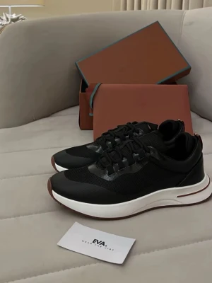 Loro Piana - Stilrena svarta sneakers med ovandel i mesh och syntet, svarta snören och vit chunky sula med brun detalj. Perfekta för en trendig och sportig look.