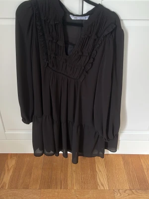 Svart volangklänning från Zara - Svart kortklänning från Zara med långa ärmar och v-ringning. Klänningen har fina volangdetaljer framtill och är tillverkad i ett lätt, flödande material som ger en luftig känsla. Perfekt för dig som gillar en stilren och trendig look.