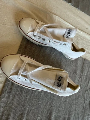 Vita Converse All Star låga sneakers - Klassiska vita Converse All Star sneakers i canvas med lågt skaft och vit gummisula. Skorna har den ikoniska loggan på plösen och detaljer i silver vid snörningen. Perfekta för en avslappnad och stilren look.