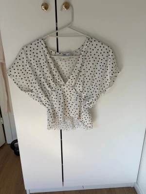 Prickig topp från Zara - Snygg vit topp från Zara med svarta prickar. Toppen har v-ringning, korta vida ärmar och smockad midja. 