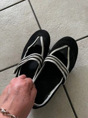 Svarta ballerinaskor med silvriga band storlek 37 - Snygga svarta ballerinaskor med unika silvriga band över foten. Skorna har rund tå och är gjorda i mockaliknande material. Perfekta för dig som vill ha en cool och stilren look. Märke ej synligt.
