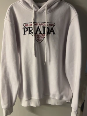 Vit hoodie från Prada med tryck - Vit hoodie från Prada med svart och rött broderat logotyptryck på bröstet. Klassisk modell med huva och dragsko samt ribbade muddar vid ärmslut och nederkant. Mjuk bomullskänsla och avslappnad passform.