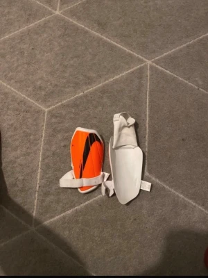 Nike benskydd orange med svart detalj - Säljer ett par benskydd från Nike i orange med svarta detaljer och vita kanter. De har vita remmar och en elastisk del för att sitta bra på benet. Perfekta för fotbollsträning eller match.