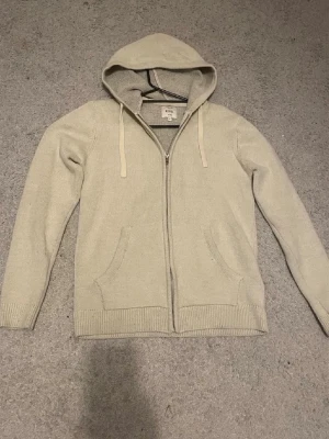 Beige stickad hoodie från Rebel - Snygg beige stickad hoodie från Rebel med dragkedja framtill och två fickor. Tröjan har huva med dragsko och ribbade muddar vid ärmslut och nederkant. Perfekt för en avslappnad och stilren look.