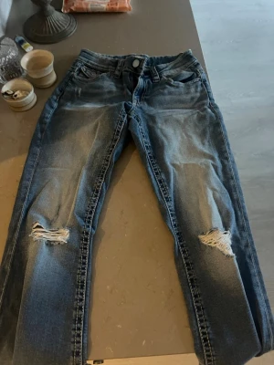 Blå slitna jeans med hål på knäna - Säljer ett par blå jeans med snygga slitningar och hål på båda knäna. Jeansen har klassisk femficksmodell, normal midja och raka ben. Perfekta för en avslappnad och trendig look. Materialet är denim i bomull.