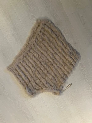 Beige kaninpäls poncho - Mysig beige poncho i kaninpäls med ribbad struktur och ett litet band för att knyta. Perfekt för att ge en lyxig touch till din outfit och hålla dig varm under kyliga dagar.