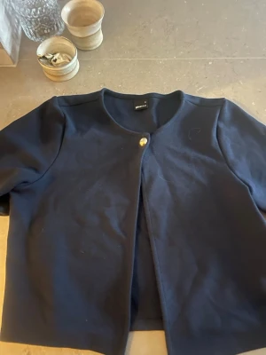 Mörkblå kofta med knapp från Gina Tricot - Stilren mörkblå kofta från Gina Tricot med rund halsringning och en dekorativ guldig knapp framtill. Koftan har kortare ärmar och är tillverkad i ett mjukt tyg som känns bekvämt mot huden. Perfekt för att lyfta en enkel outfit.