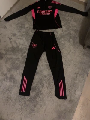 Arsenal träningsset svart/rosa Adidas  - Säljer ett Arsenal träningsset från Adidas i svart med rosa detaljer. Setet består av en långärmad tröja och matchande byxor, båda med klubbmärke och Adidas-logga. Materialet är troligtvis polyester, perfekt för träning eller uppvärmning. Snygg design med rosa ränder och tryck.