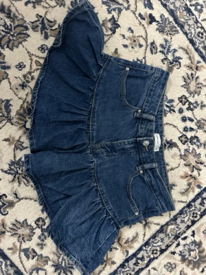 Blå volangkjol i denim från Gina Tricot - Kort jeanskjol från Gina Tricot i klassisk blå denim. Kjolen har volang nedtill, femficksmodell och bälteshällor i midjan. Perfekt för dig som gillar en trendig och avslappnad stil. Stängs med knapp och dragkedja framtill.