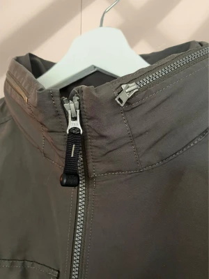 Grå vindjacka från Jack Wolfskin - Snygg grå vindjacka från Jack Wolfskin med dragkedja framtill, två bröstfickor med knappar och två snedställda fickor med dragkedja. Jackan har hög krage, huva och diskret logga på bröstet samt ärmen. Perfekt för blåsiga dagar.