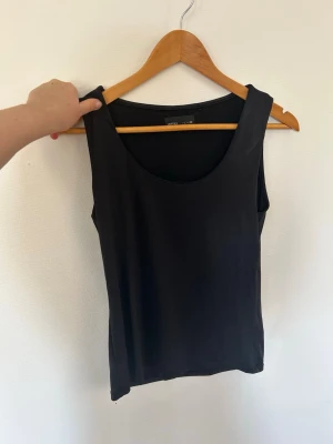 Svart linne från Vero Moda - Enkel och stilren svart ärmlös topp från Leftise med rund halsringning. Tillverkad i ett mjukt och stretchigt material som sitter skönt mot kroppen. Perfekt att matcha med jeans eller kjol för en clean look.