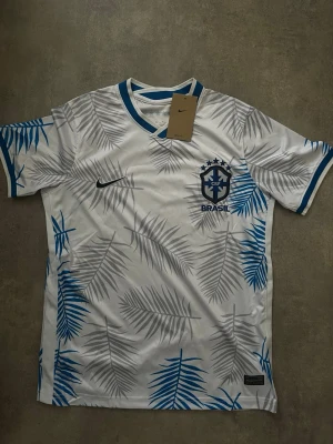 Vit Brasilien fotbollströja Nike - Snygg vit fotbollströja från Nike med Brasilien-logga och blå Nike Swoosh. Tröjan har grå och blå palmbladsmönster samt blå detaljer vid krage och ärmslut. Perfekt för dig som älskar fotboll och vill sticka ut på planen.