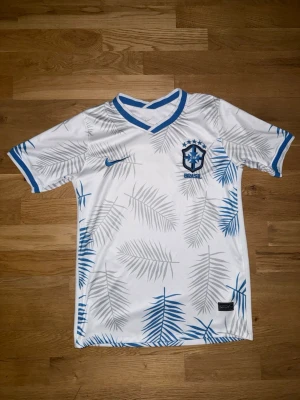 Vit Brasilien fotbollströja Nike - Snygg vit fotbollströja från Nike med Brasilien-emblem och blå detaljer. Tröjan har blå och grå palmbladsmönster, blå Nike-logga och blå ribbade kanter vid ärmar och krage. Perfekt för dig som älskar fotboll och vill sticka ut på planen.