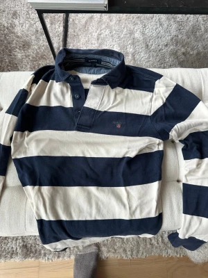 Randig rugbytröja från Gant - Snygg långärmad rugbytröja från Gant med breda marinblå och vita ränder. Klassisk krage och knäppning framtill. Perfekt för en sportig och avslappnad stil.