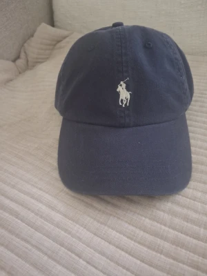 Mörkblå keps från Polo Ralph Lauren - Snygg mörkblå keps från Polo Ralph Lauren med klassisk böjd skärm och broderad logga framtill. Justerbar rem baktill för perfekt passform. Tillverkad i bomull för en bekväm känsla och stilren look.