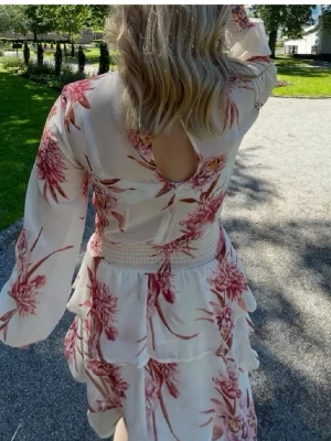 Blommig volangklänning från Gina Tricot - Säljer en vit klänning med rosa blommor från Gina Tricot. Klänningen har långa ärmar, smock i midjan och volangdetaljer på kjolen. 