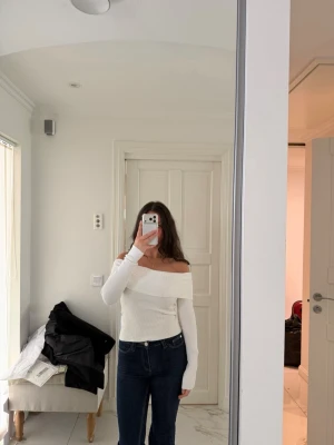 Vit offshoulder ribbad topp - Trendig vit offshoulder topp med ribbad struktur och långa ärmar. Sitter tight och har en bred kant över axlarna som ger en snygg siluett. Perfekt att styla med jeans eller kjol för en clean look.