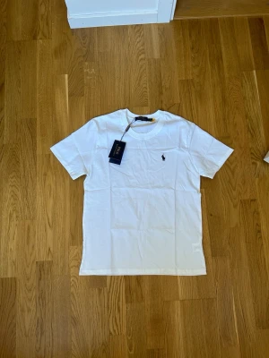 Vit t-shirt från Polo Ralph Lauren - Klassisk vit t-shirt från Polo Ralph Lauren med kort ärm och rund halsringning. Liten broderad logga på bröstet. Perfekt basic-plagg i mjuk bomull som passar till allt.