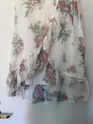 Blommig omlottkjol med volang - Söt omlottkjol i vitt med blommigt mönster i rosa och grönt. Kjolen har volangkant och knytband i midjan, vilket ger en romantisk och somrig känsla. Perfekt för dig som gillar en feminin och luftig stil.